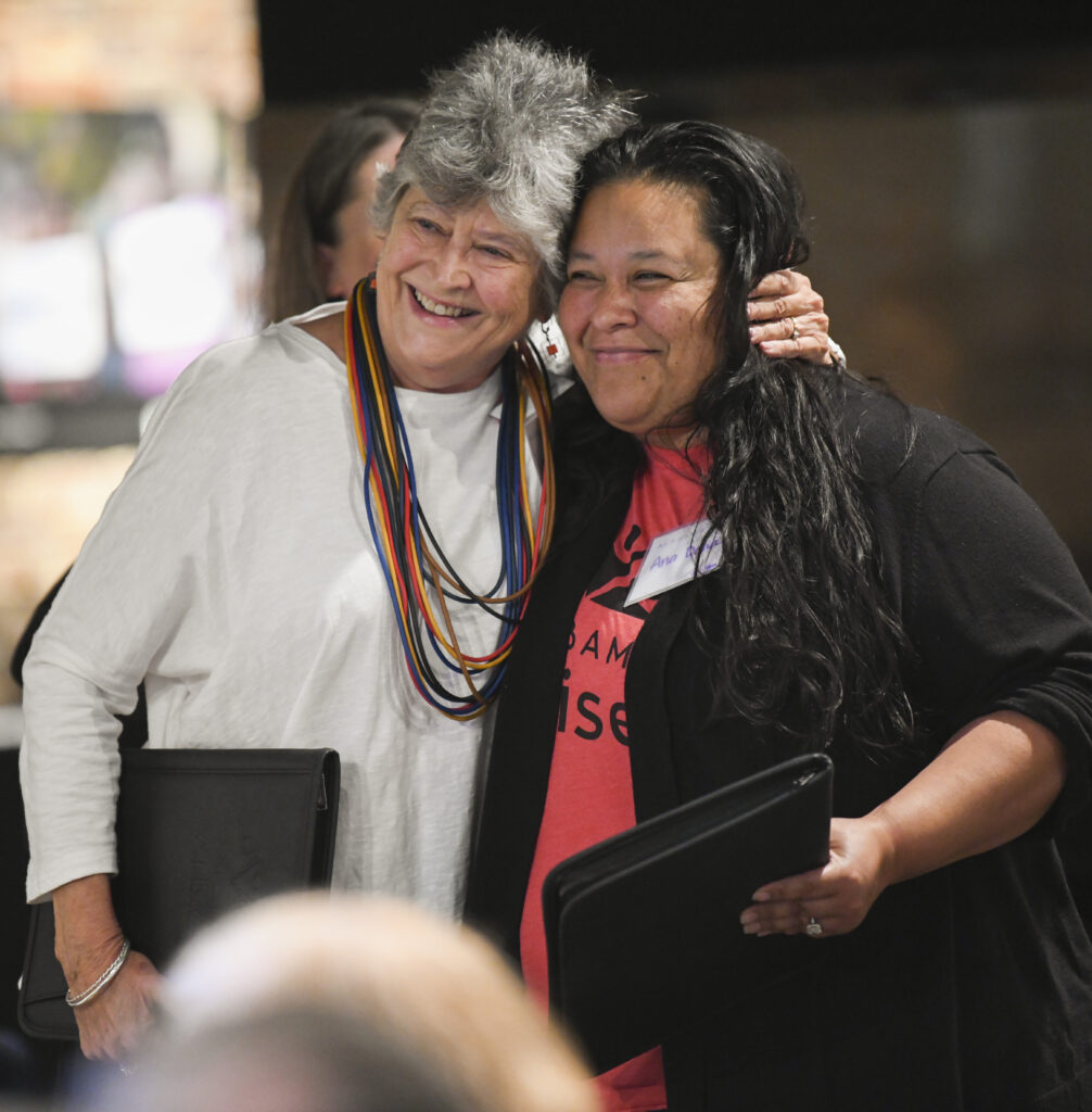 Outgoing Alabama Arise board president Kathy Vincent, a white woman with a white shirt, hugs outgoing Alabama Arise board member Ana Delia Espino, a Hispanic woman with a red shirt and a black sweater.