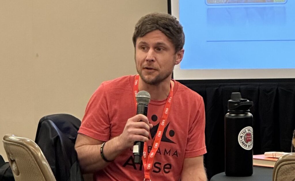 A white man with dark hair holds a microphone. He is wearing a light red shirt with a black Alabama Arise logo.