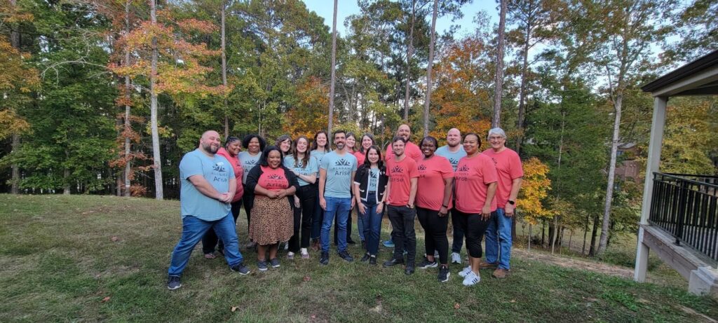 A group of 17 people smile for a photo while standing on a green hilltop. All are wearing red or green T-shirts with the Alabama Arise logo. Behind them is a line of evergreen trees and trees with leaves turning yellow or orange for fall.