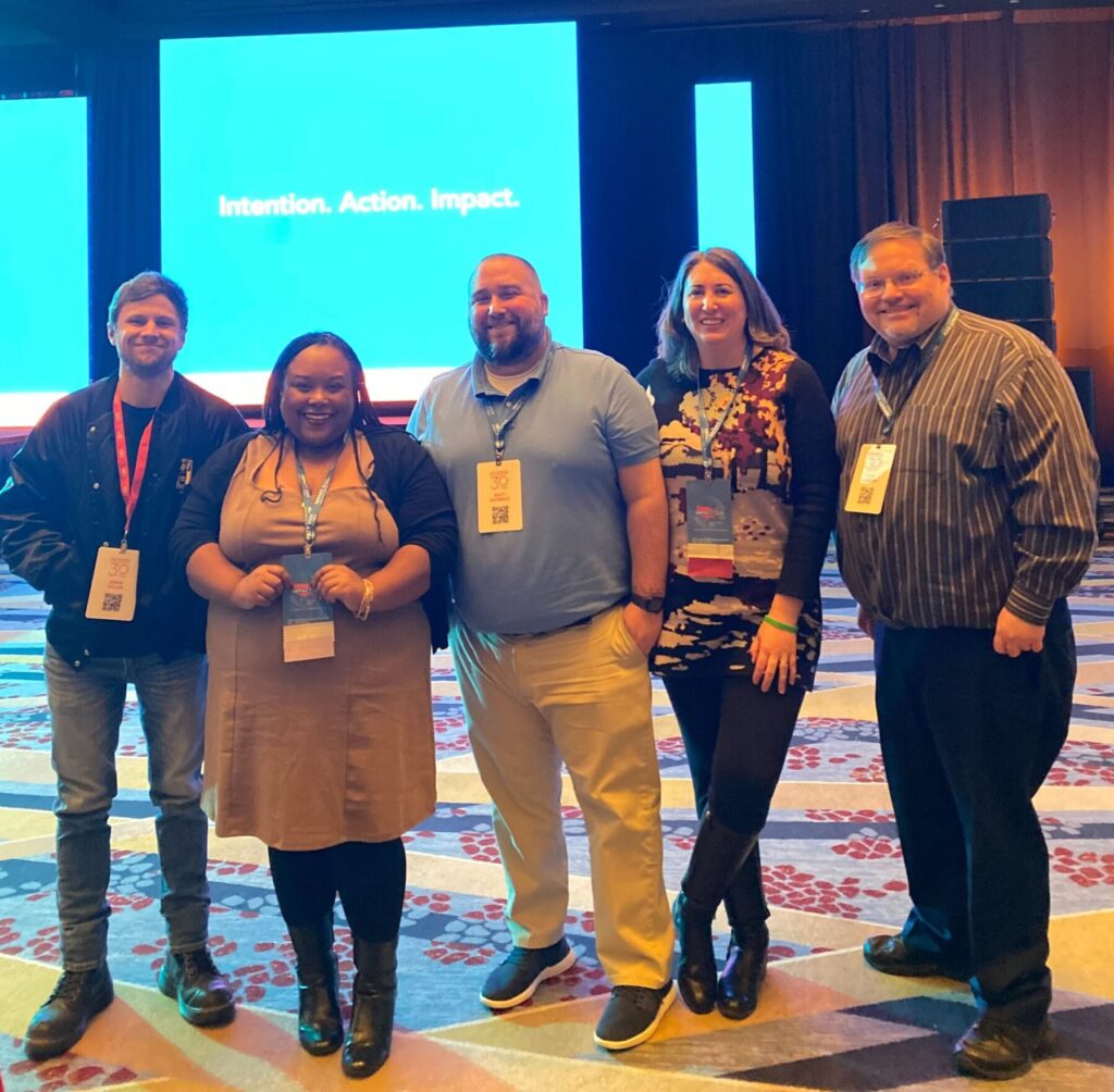 A white man in a black jacket, a Black woman in a light brown dress, a white man in a light blue shirt, a white woman in a red and black blouse, and a white man in a dark blue shirt stand and smile. Behind them is a blue screen with white text reading "Intention. Action. Impact."