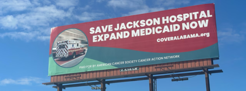 A red and green billboard with a picture of an ambulance and white text reading "Save Jackson Hospital. Expand Medicaid Now." The billboard directs viewers to coveralabama.org.