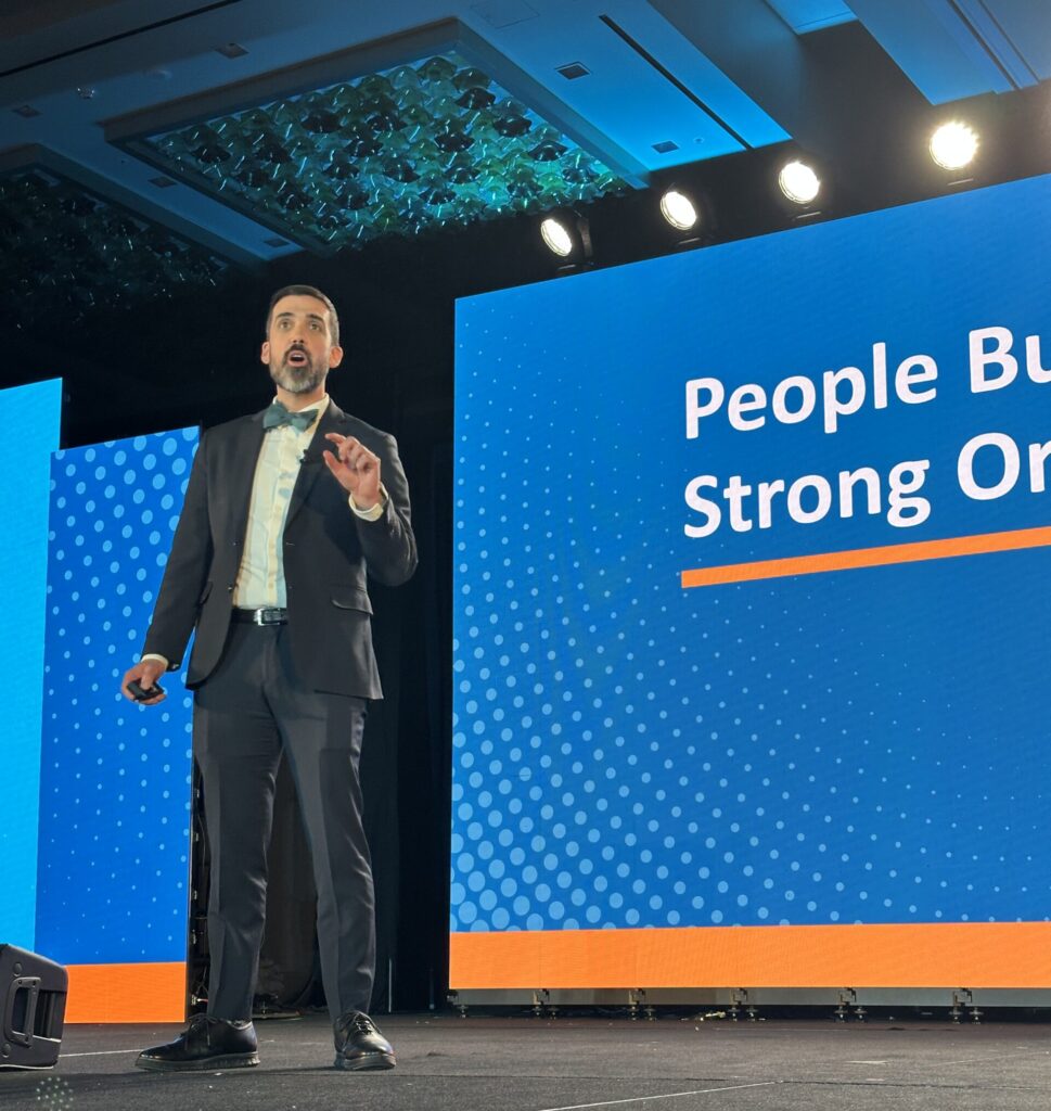 A white man with dark hair and a beard speaks in front of blue screen with white text reading "People Build Strong Organizations." He is wearing a black suit and a bowtie.