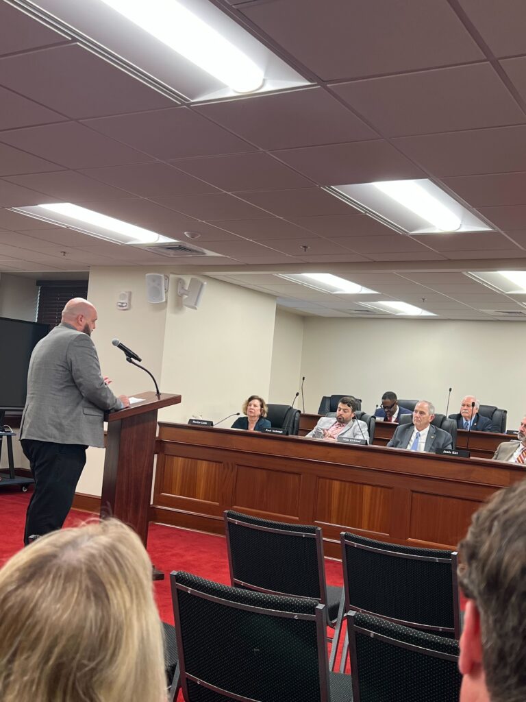 A man stands at a podium speaking to lawmakers who are seated.
