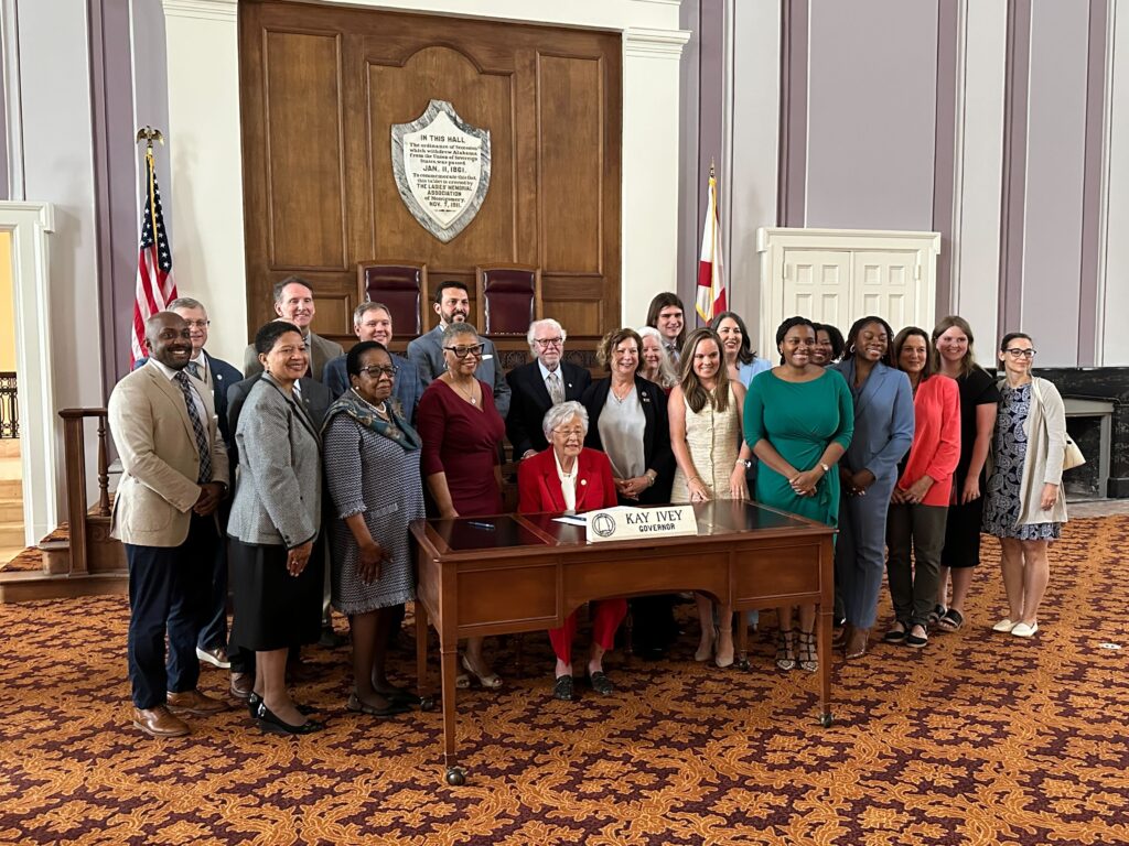 Gov. Kay Ivey sits behind a wooden desk with her nameplate on it and U.S. and Alabama flags in the background. Smiling people stand behind and to both sides of her. Behind her is wooden paneling and walls with a gray and white pattern. The patterned carpet is dark red and orange.