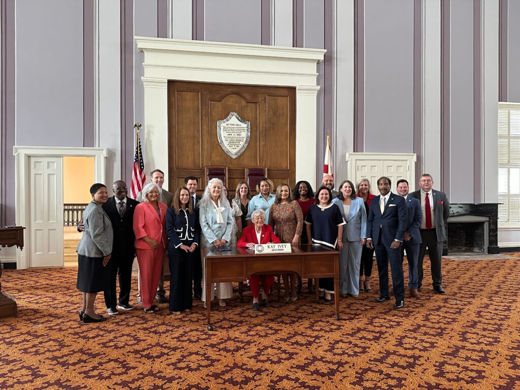 Gov. Kay Ivey sits behind a wooden desk with her nameplate on it and U.S. and Alabama flags in the background. Smiling people stand behind and to both sides of her. Behind her is wooden paneling and walls with a gray and white pattern. The patterned carpet is dark red and orange.