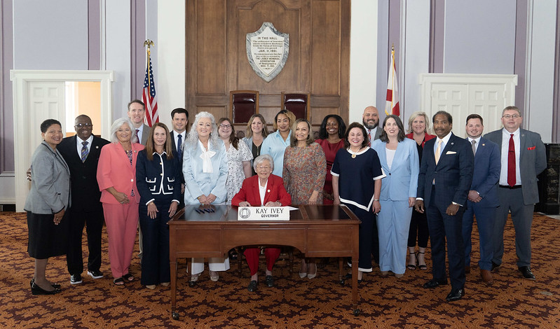 Gov. Kay Ivey sits behind a wooden desk with her nameplate on it and U.S. and Alabama flags in the background. Smiling people stand behind and to both sides of her. Behind her is wooden paneling and walls with a gray and white pattern. The patterned carpet is dark red and orange.