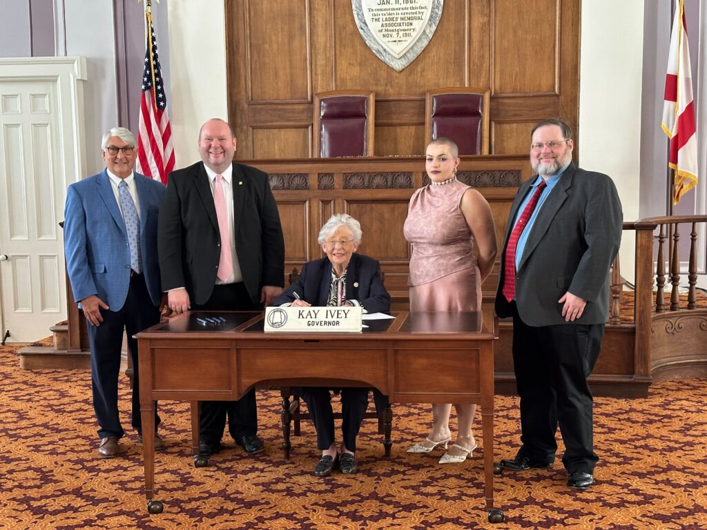 Three men wearing suits and ties and a woman wearing a pink dress stand to either side of Gov. Kay Ivey, who is wearing a black jacket and seated behind a wooden table.