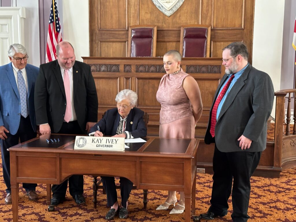 Three men wearing suits and ties and a woman wearing a pink dress stand to either side of Gov. Kay Ivey while she signs a bill. Ivey is wearing a black jacket and seated behind a wooden table.