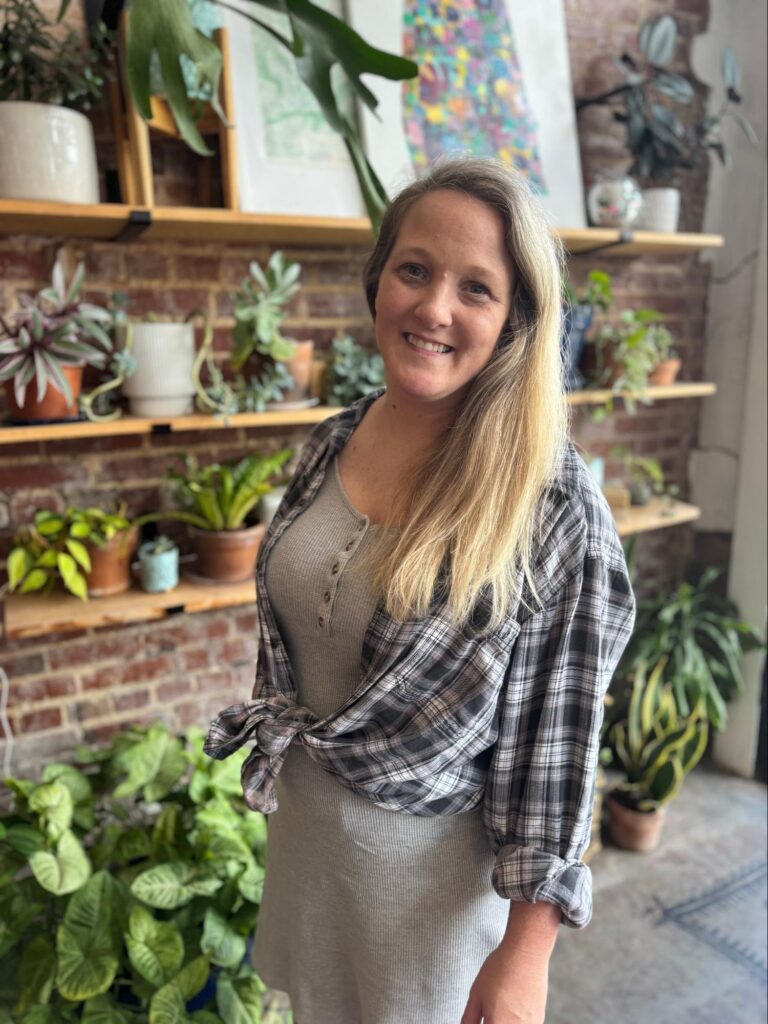 An adult woman stands in front of a wall of plants.