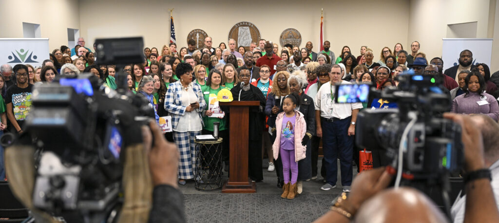 Dozens of people stand behind and to either side of a lectern in a committee room at the Alabama State House. Close-up shots of two videographers are in the foreground.