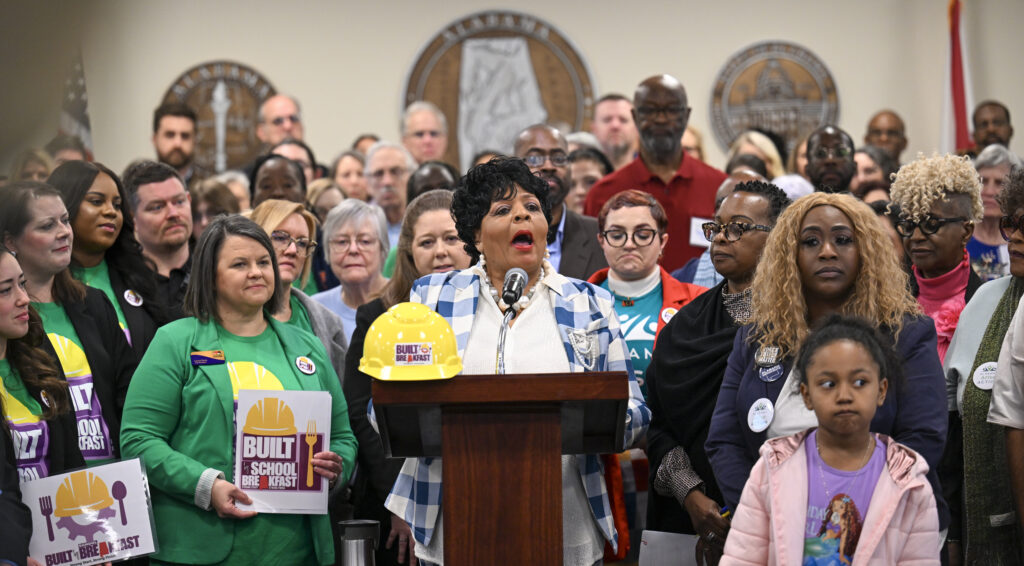 A woman speaks behind a lectern in a committee room at the Alabama State House. Dozens of people stand behind and beside her.