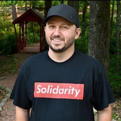 A smiling man in a black shirt and black hat.