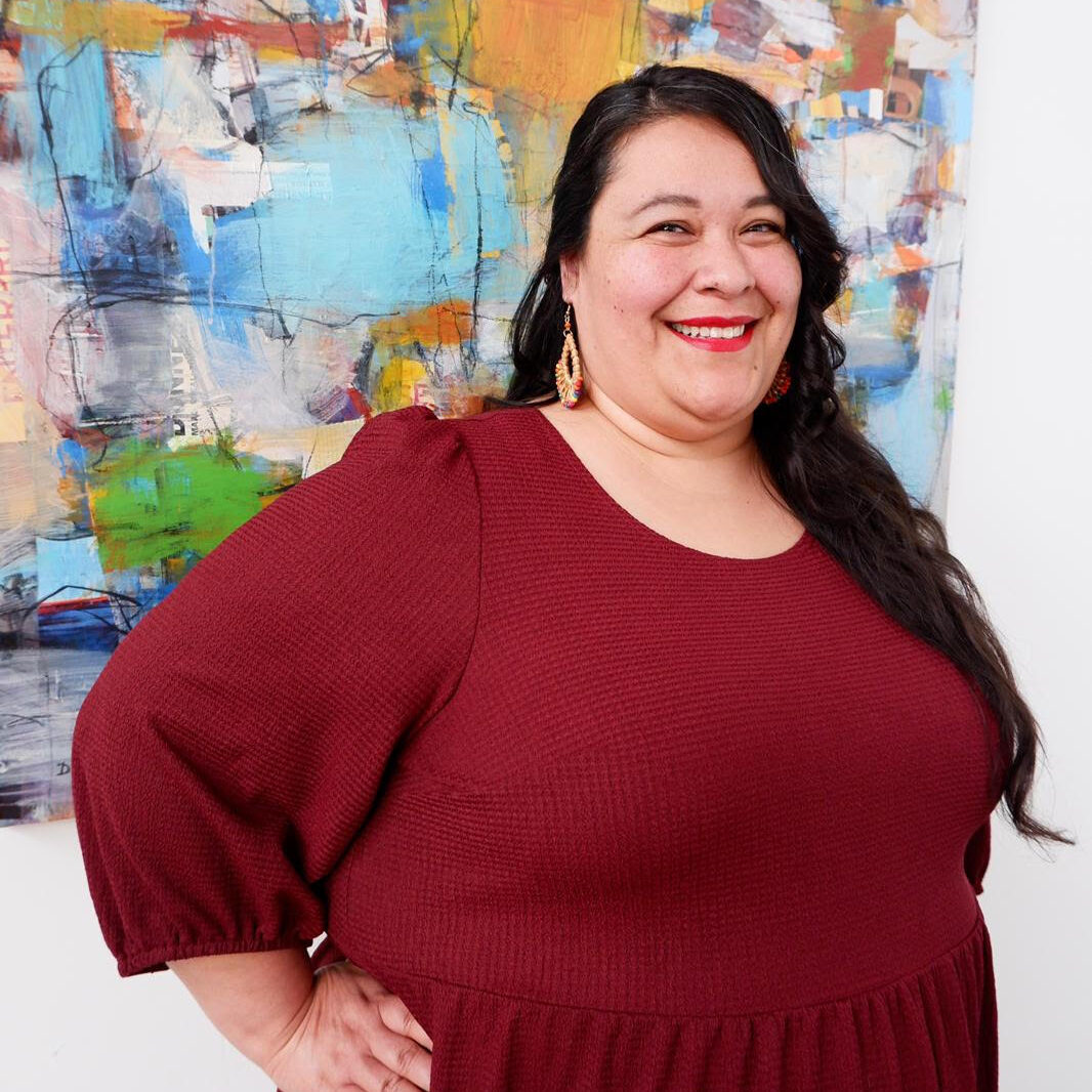A smiling Latina with long dark hair and wearing a red dress stands in front of a colorful painting.